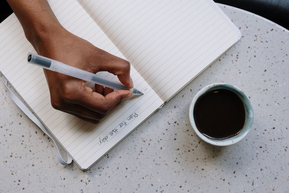 A hand journaling 'Plan for the day' beside a cup of coffee